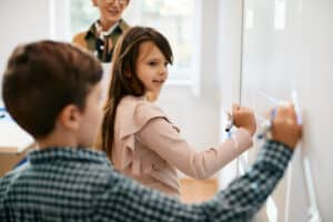 Schoolgirl and her classmate writing on a board in an elementary school with a happy teacher in the background. Student success and retention concept