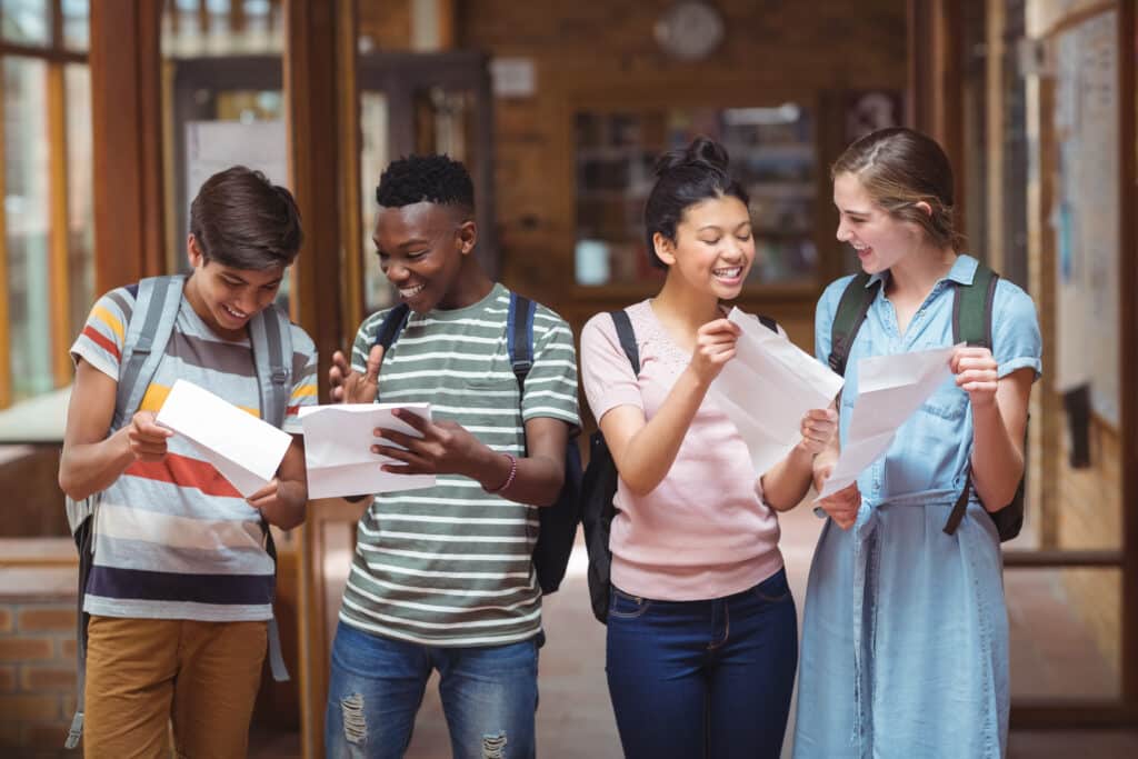 Happy classmates looking at grade cards in corridor at school